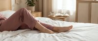 Woman relaxing in comfortable pink pajamas on white bedding in bright modern bedroom with natural morning light streaming through window.