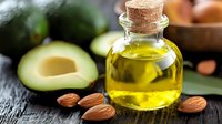 Fresh avocado oil in glass bottle with cork and raw almonds on dark wooden background next to cut ripe avocado, healthy organic cooking ingredients.