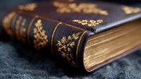 Antique leather-bound book with ornate gold floral embossing on dark spine, lying on textured black surface. Aged pages visible showing vintage patina.