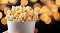 Fresh popcorn overflowing in white paper container against blurred warm bokeh lights background, creating festive cinema and entertainment atmosphere.