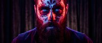 Bearded man with ornate skull face paint in red and blue colors for Day of the Dead celebration against dark background with dramatic lighting effects.