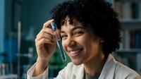 Young African American female scientist in lab coat holding test tube with blue liquid, smiling while conducting research in modern laboratory setting.