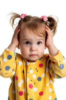 Toddler girl in yellow polka dot dress with pigtails and red checkered bows holds hands to head with surprised expression against white background.