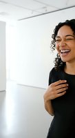 Young woman with curly dark hair and medium skin tone wearing black shirt, laughing with eyes closed in bright modern interior space for lifestyle and emotion content.