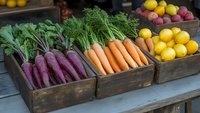 Fresh organic purple carrots and traditional orange carrots displayed in rustic wooden crates alongside colorful citrus fruits at farmers market produce stand.