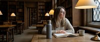 Young blonde woman studying with water bottle in historic university library with wooden shelves and warm lighting creating peaceful academic atmosphere.