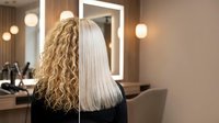Woman showing hair transformation from curly to straight blonde in modern salon with professional styling equipment and mirror lighting.