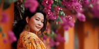 Young Hispanic woman in traditional golden embroidered dress smiling among vibrant pink bougainvillea flowers in warm sunlight for cultural celebration.