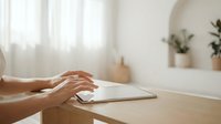 Woman hands typing on white tablet computer at wooden desk in bright modern home office with natural light and plants in background.