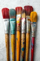 Colorful used paintbrushes with wooden handles and paint splatters arranged in row on white textured background, showing artistic wear and creative process.