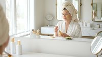 Young woman in white bathrobe and towel applying skincare cream in modern bathroom mirror reflection morning routine self-care wellness spa treatment.