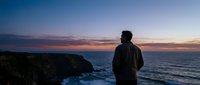 Man in dark jacket standing on coastal cliff overlooking ocean at sunset, contemplative mood, suitable for travel, lifestyle, and inspirational content.