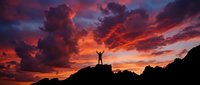 Silhouette of person with raised arms celebrating success on mountain peak against dramatic orange and purple sunset sky with storm clouds.