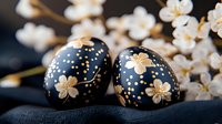 Black Easter eggs decorated with golden cherry blossom pattern and dots, placed near blooming white spring flowers on dark background, festive holiday composition.