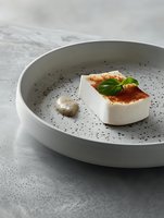 Modern plated dessert featuring white panna cotta with dusted spices and fresh basil leaf, served on textured gray ceramic plate against concrete background.