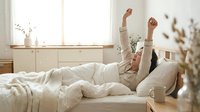Young woman stretching arms up while lying in bed during sunny morning, expressing joy and energy after good night sleep in cozy bedroom interior.