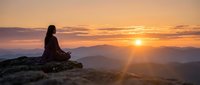 Woman meditating on mountain peak during golden sunrise with layered hills and warm sky colors creating peaceful spiritual moment for wellness and mindfulness.