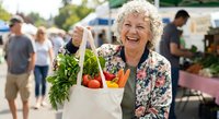 Happy senior Caucasian woman holding eco-friendly shopping bag with fresh vegetables at farmers market. Sustainable lifestyle and healthy eating concept.