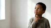 Young boy with dark skin tone and black hair wearing a beige ribbed sweater, hand to chin in thoughtful pose, looking upward against neutral white wall with natural light.