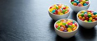 Colorful candy-coated chocolates in white ceramic bowls arranged on dark slate surface for confectionery and sweet treat marketing materials.