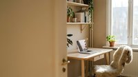 Minimalist home office workspace with wooden desk, laptop, potted plant, floating shelves, and comfortable chair by bright window with natural light for remote work and productivity.
