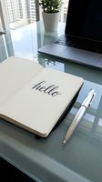 Open notebook with hello lettering beside a silver pen on a glass desk, laptop and plant in background, modern workspace.