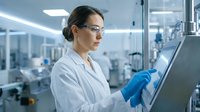 Female scientist in white lab coat and safety glasses working on computer in modern laboratory for research and development.