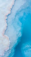 Aerial view of natural salt lake shoreline with turquoise water meeting white crystalline deposits, creating abstract patterns and gradients in blue tones.