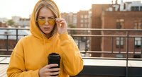 Young blonde Caucasian woman in yellow hoodie and sunglasses holding coffee cup on urban rooftop terrace during golden hour sunset with city buildings.