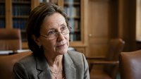Mature woman with shoulder-length brown hair and glasses wearing gray blazer in professional library setting with wooden furniture and bookshelves, suitable for business, education, and corporate communications.