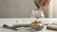 Hand dropping golden coins into glass jar on marble surface with newspaper and notebook for savings and financial planning concept.