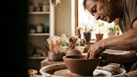 Senior male potter crafting ceramic bowl on spinning wheel in warm sunlit workshop studio with shelves of pottery tools and finished pieces in background.