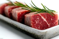 Raw beef cubes with fresh rosemary sprigs arranged on stone plate, showing marbled texture and rich red color. Close up view highlights premium meat quality.