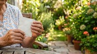 Senior woman holding tablet device while sitting outdoors in garden with flowering plants and greenery in background.