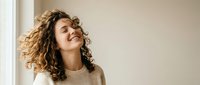 Young woman with curly hair laughing joyfully in cozy sweater by window, expressing happiness and positive emotions in warm natural light.