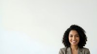 Young woman with dark curly hair and olive complexion wearing a neutral blazer, smiling at camera against white background for corporate portraits and professional headshots.