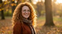 Young Caucasian woman with curly red hair and glasses smiling warmly in autumn park during golden hour sunset light wearing cozy brown sweater and scarf.