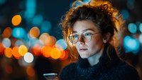 Young woman with curly hair and glasses in urban night setting with colorful bokeh lights creating atmospheric portrait for lifestyle content.