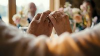 Newlywed couple holding hands with wedding rings during ceremony celebration with guests and flowers in background.