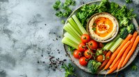 Fresh hummus dip served with raw vegetables including carrots, celery, broccoli and cherry tomatoes on vine. Mediterranean appetizer platter on gray background.