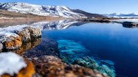 Tranquil alpine lake with crystal clear blue water showing underwater rocks, surrounded by snow-capped mountains reflecting on the calm surface.
