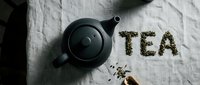 Black ceramic teapot on linen cloth with loose green tea leaves arranged to spell TEA, flat lay composition for tea brand or lifestyle blog.