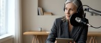 Professional middle-aged Caucasian woman podcaster recording audio content in modern home studio setup with microphone and laptop for broadcasting.
