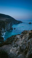 Hiker standing on rocky cliff overlooking turquoise ocean waters and dramatic coastal cliffs at dusk, perfect for travel and adventure themes.