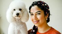 Young woman with dark hair and brown eyes, wearing an orange top, smiling while posing with a white standard poodle against a neutral background for pet and lifestyle content.