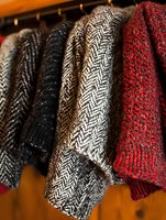 Wool tweed coats in herringbone pattern hanging on wooden rack, featuring classic black, gray and red colors, showing texture and weave detail in close-up view.