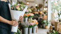 Florist holding wrapped bouquet of mixed flowers in shop with blurred floral arrangements in background, suitable for flower business, retail, and floristry concepts.