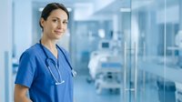 Confident female healthcare professional in blue scrubs with stethoscope standing in modern hospital corridor with glass walls and medical equipment.