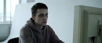 Young Caucasian male programmer in hoodie working at computer desk in modern office environment with natural lighting from window.