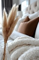 Dried pampas grass in glass vase against cozy bedroom setting with soft white blanket, beige pillows and tufted headboard creating peaceful home atmosphere.